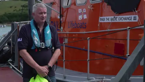 Simon Linsley stood on the left next to a large orange lifeboat. He is wearing a blue float around his neck, a blue RNLI t-shirt and is holding a yellow item. 