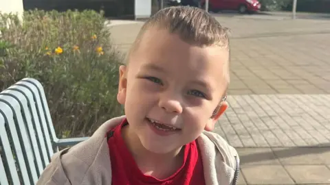 A small boy smiling in a red shirt and hoodie smiles while sitting on a bench. He has a device attached to his ear.