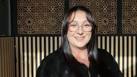 Vikki Irwin/BBC Corrin Wallace is sitting down in a room with a wooden patterned cladding on the walls. Corrin is wearing a black jacket and glasses and is smiling at the camera 