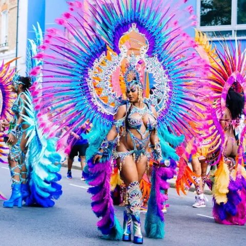 @draculaxproductions Rachel Alexander is seen in a vibrant feathered costume with bold colours and jewels leads dancers in a carnival parade, wearing elaborate wings, a jewelled headpiece, and platform boots.