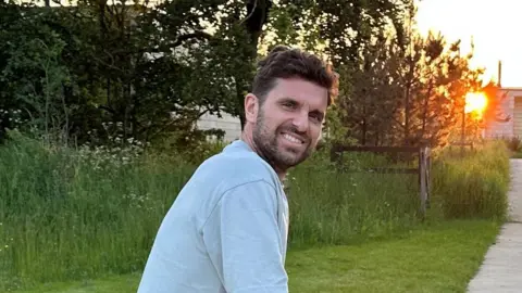 Theo Schooling stood outside as the sun is setting. There is long grass and trees in the background. He has brown hair and beard.