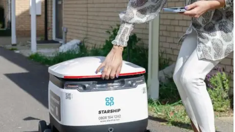 Cambs County Council Woman reaches out to lift robot lid to remove a delivery. The robot is on a residential street. The woman holds a mobile phone in one hand. 