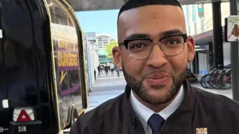 Martin Heath/BBC Mason Humberstone with very short dark hair and glasses, smiling at the camera and wearing a black jacket with a Union Jack badge. He is standing by a burger van in Stevenage town centre.