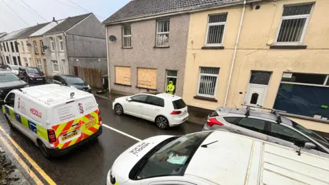 Police officer stands in doorway of partially boarded up house. Police van nearby.