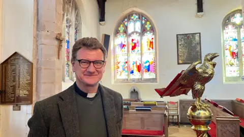 Kerry Devine Reverend Canon Mark Collinson has brown wavy hair and glasses, he has a tweed jacket with a dark green 'v' necked jumper with his dog collar beneath.
To his right is a gold eagle lecturn with pews visible too and a stained glass window.
