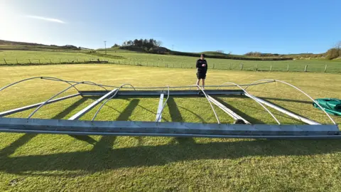 A grey metal frame is lying on a green field. It looks like it has been bent out of shape. There is a blue sky in the background and a person in a black top and shorts is stood behind the frame.