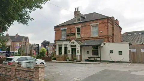 Streetview image of the Sportsman pub, a substantial brick Victorian building standing along in an urban setting with parking area in front