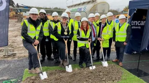 Greater Manchester Mayor Andy Burnham and other local dignitaries, all wearing white hard hats and yellow hi-vis jackets, pose with spades on a building site.