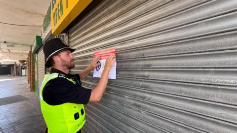 A police officer in short black shirt, helmet and high-vis jacket is seen posting a closure order on the door of the Cradley market shop