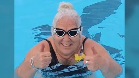 John Devine/BBC A woman in  a swimming pool wearing dark swimming goggles faces the cameras and raises two thumbs up.