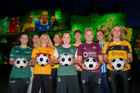 PA Media Two rows of female footballers standing in front of Edinburgh Castle, which is lit up by a green projection. The women are all wearing football strips and those in the front row are holding footballs.