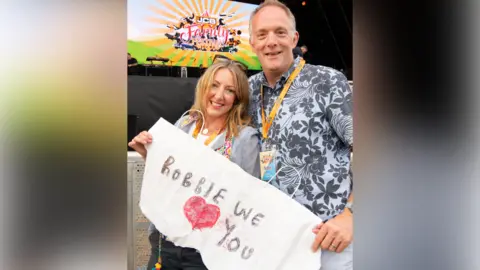 JCB A woman and a man stand at the barrier in front of a stage while holding a white banner which says "Robbie we love you" - the "love" is represented with an image of a red heart.