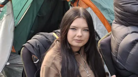 A young woman with long brown hair and wearing a brown jumper sits on a camping chair surrounded by tents. 
