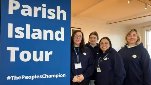 Four women from Citizen's Advice Jersey are smiling at the camera. They are wearing navy branded hoodies and lanyards and are standing next to a big pop-up bright blue banner with white lettering that reads Parish Island Tour #ThePeoplesChampion