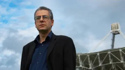 Elizabeth Weiss A man wearing a navy shirt and a black coat with black round-framed glasses. Behind him is what looks like a radio telescope. 