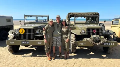 Two World War Two military jeeps parked in a beach car park. A man is seen with his two daughters. All are dressed in green military gear