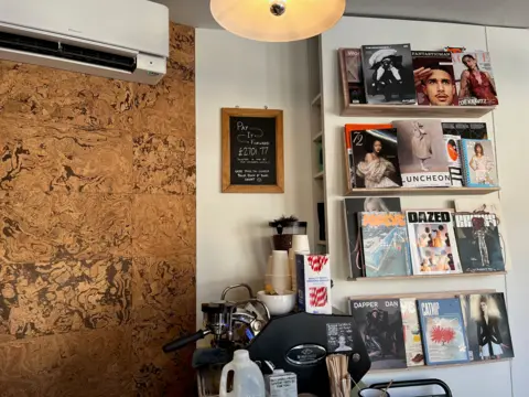 The interior of a book store with a display of magazines to the right, a coffee machine in the centre, and a black chalkboard sign that reads: Pay It Forward, £2701.77 donated so far by our wonderful community. Use this to cover your book if you're short. 