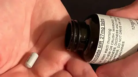 BBC A pill pictured in the palm of a hand, alongside an open medicine bottle from which the pill appears to have been tipped.