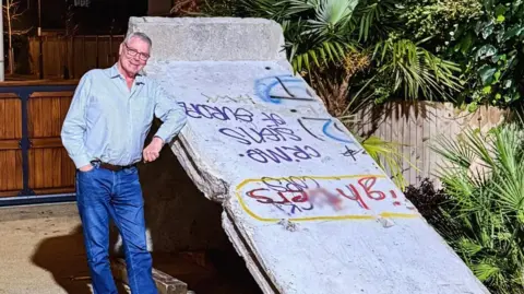 Steven Thorpe - a man in his sixties with white hair and glasses, wearing blue jeans and a blue shirt - stands next to a large section of grey wall with graffiti on it.