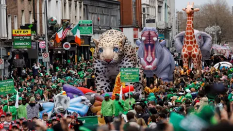 REUTERS/Clodagh Kilcoyne Large floats depicting wild animals including a leopard, hippo, giraffe and and elephant, parade through a street in Dublin.  The street is packed with people wearing green who are lining the route. 