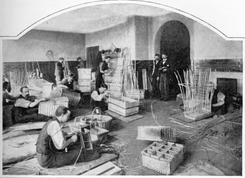 Getty Images A black and white photograph showing blind men in waistcoats sat on the floor a factory warehouse weaving baskets. Others are stood in the distance working on the baskets. Two men in three piece suits stand in an archway looking at the work.