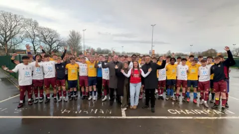 A large group of football players pose for a picture in a car park. In front of the group are several people in smart clothing and a woman in jeans and an Arsenal football shirt. Henry Nowak's dad Mark waves at the camera. 
