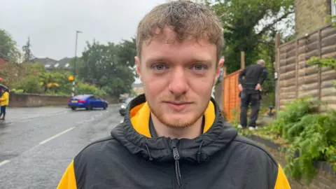 A man wearing a black and yellow coat stood in the street while it is raining. He is looking into the camera.