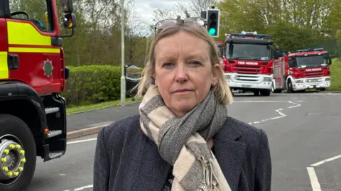 A woman with blonde hair and glasses on the top of her head in a dark blue jacket and grey and white scarf in the street with three fire engines behind her