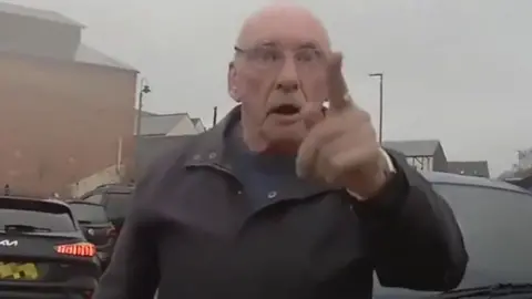A man standing in a car park. He has his finger pointed at the person wearing the body-cam.