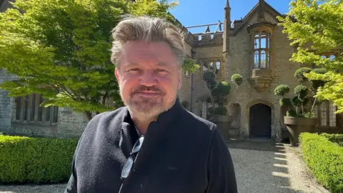 James Perkins smiles outside Parnham House. He has a beard and light brown and grey hair in a quiff and wears a black top.