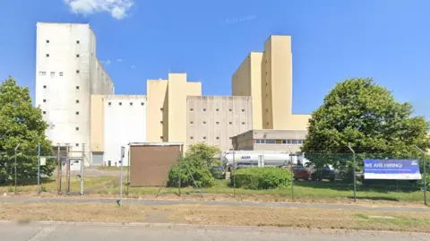Large concrete building with several storeys. There are towers on either side with a shorter section in the middle. There is a large number of small square windows on the building.  The letters ADM are visible on a smaller white building in the foreground. There are trees either side of the building.