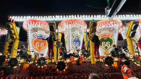 A carnival float with a biker theme. It features several performers wearing leather jackets on motorcycles and Route 66 road signs. 