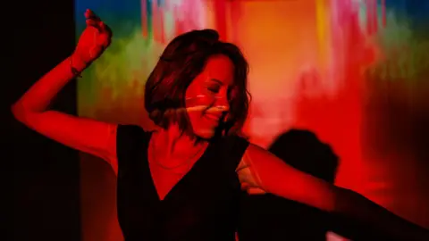 Stock image of Young woman dancing joyfully in vibrant colorful lighting, expressing excitement and energy. The dynamic lighting effects create a lively and creative atmosphere, symbolizing self-expression, freedom, and celebration.