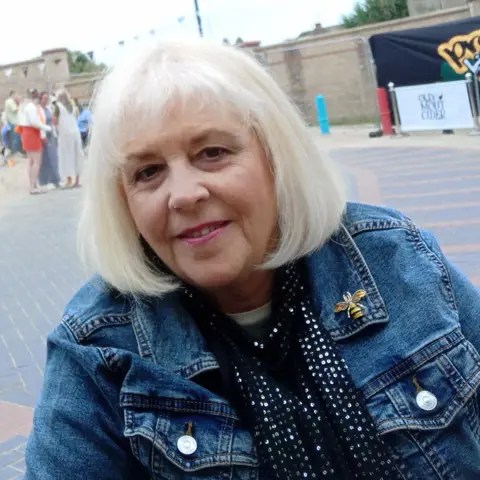Karen Beattie Karen has white hair that is shoulder length. She is smiling at the camera.
She is wearing a denim jacket with a bee as a broach on the right collar of the jacket. Karen has a black and silver scarf around her neck.