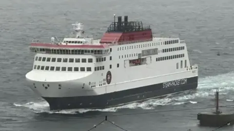 The Manxman ferry, a tall white ship, with red detail on the top and dark base, driving through waves at sea. A buoy can be seen on the right and some festoon street lighting in the close foreground on a grey da y.
