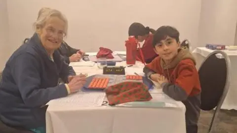 ABSP Diane has her silver hair tied up, and wears glasses and a dark blue jacket. Jake has short black hair and wears a red and blue top with a brown hood. They sit opposite each other at a table and smile at the camera, a Scrabble board in between them.