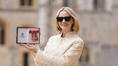 Carey Mulligan holds up a CBE medal in a box while posing for a photo in the grounds of Windsor Castle. She has a blonde bob, and is wearing sunglasses and a cream embroidered jacket.
