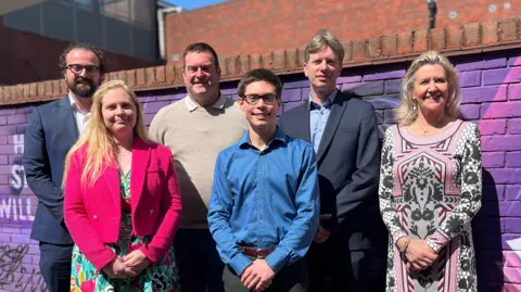 Six smartly-dressed candidates for Huntingdonshire District Council elections pose for a photograph. Two woman and one man are on the front row, and three men are on the back row. They are stood in front of a purple graffiti-d wall.