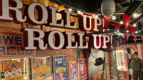 A large sign saying 'roll up roll up' over an exhibition of circus posters at the museum