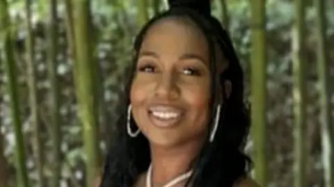 A young woman wearing gold jewellery smiles. She is standing in front of a forest.