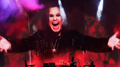 An huge picture of Ozzy Osbourne, with his mouth wide open in his trademark snarl, wearing black and his arms outstretched. The image is bathed in smoke and red and purple lights above musicians performing on stage.
