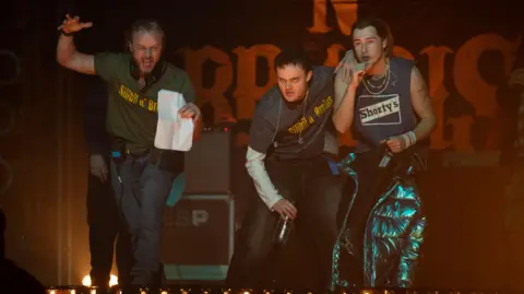 Glasgow Film Festival James McAvoy - wearing a green T-shirt saying Silibil N' Brains and holding a piece of paper in one hand, gestures to Séamus McLean Ross and Samuel Bottomley (in character as the rappers) with direction during filming of California Schemin'. 
