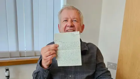 A man with short light hair sits for the camera while holding up a piece of paper with blue handwriting on it. He wears a dark blue shirt with a light blue checked pattern on it. Behind him is a window with vertical blinds.