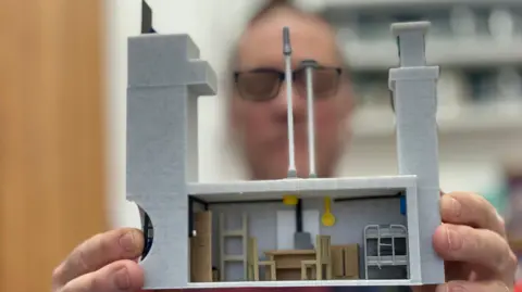 A man, whose face is out of focus, is holding a plastic scale-model of a bunker. It shows an underground room with a table, two chairs and a bunk bed. On the left, a large tower offers access to the bunker. On the right a chimney, of the same height, offers ventilation. 