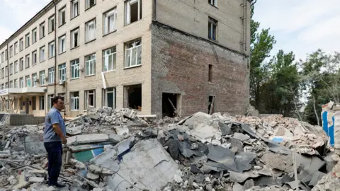 Reuters A man in a blue shirt and black trousers standing on the rubble of a fallen building, with another block of flats in the background