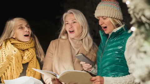 Three women singing outdoors at night wrapped in winter clothes holding sheet music