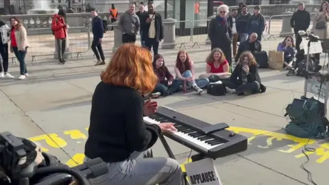 A busker playing an electric keyboard to a seated crowd in Trafalgar Square, with passers-by standing behind temporary barriers nearby.