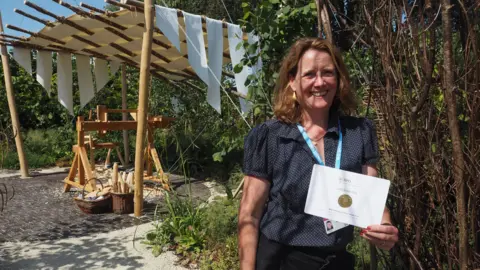 Emma Bradshaw Rachael Austin smiles as she holds a certificate bearing a small gold circle in front of her award-winning garden. There is a workshop area behind her, shaded by large wooden poles with strips of white fabric woven through them. 