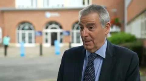 BBC Prof David Green who has short grey/black hair and wears a blue suit over a light blue shirt with a blue tie which has white spots on it. He stands in front of a red brick building with arched windows.