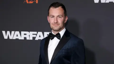 Getty Images Aaron Mackenzie in a navy tuxedo stands next to a Warfare branded wall, posing for photos at a film premiere.